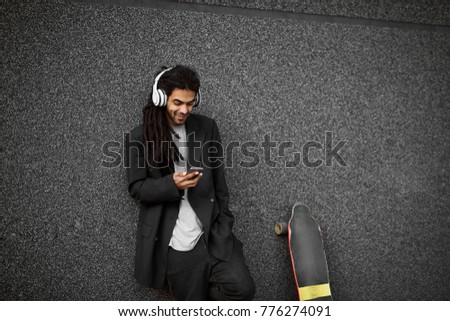 Stylish handsome bearded dreadlocks hipster skater man in a suit standing leaned on the grey wall and holding mobile in one hand while listening music with skate next to him.