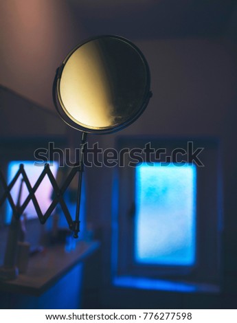 Magnifying mirror in dark eerie bathroom with small window.