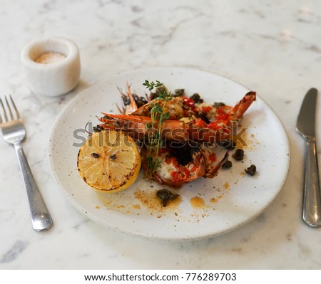 Garlic Tiger prawns with brown butter capers sauce served on white plate set on white marble top table with fork and knife and a jar of salt