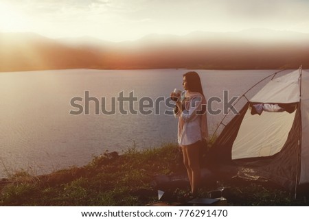 Pretty asian young woman with hot tasty drink spending vacations among stunning morning light summer landscape.
