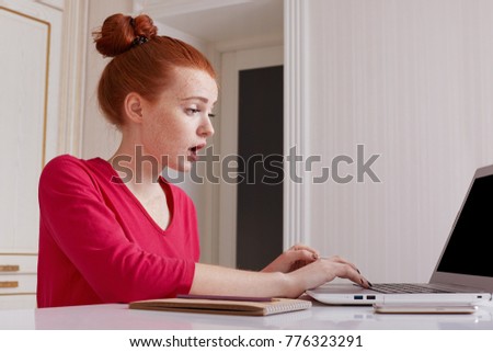 Puzzled female talented editor of online issue works remotely at home, dressed casually, has red hair tied in hair bun, creats article, sits in front of laptop at workplace with diary for making notes