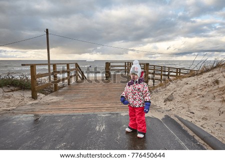 Little girl wearing jacket and red pants and hat is playing at the beach near baltic sea on cloudy sky in winter time