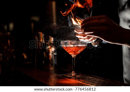 Bartender`s hands sprinkling the juice into the cocktail glass filled with alcoholic drink and making a smoky note on the dark background Royalty-Free Stock Photo #776456812