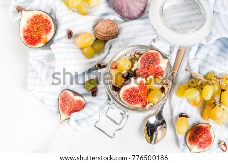 Autumn breakfast ideas, recipes. Jar of overnight autumn oats with red figs, grapes and walnuts. On white marble table, copy space top view