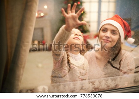 Adorable little girl wearing knitted sweater having fun with pretty mother while gathered together at cozy living room decorated for Christmas celebration and drawing picture on frozen window