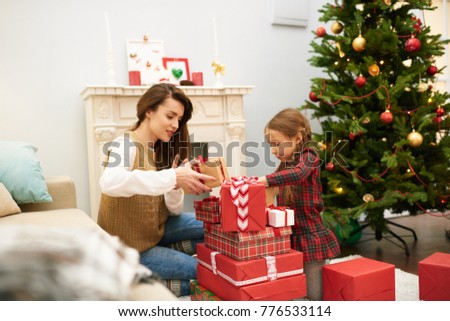 Loving family of two gathered together at cozy living room decorated for Christmas celebration and unwrapping gift boxes with interest, Christmas tree and fireplace behind them