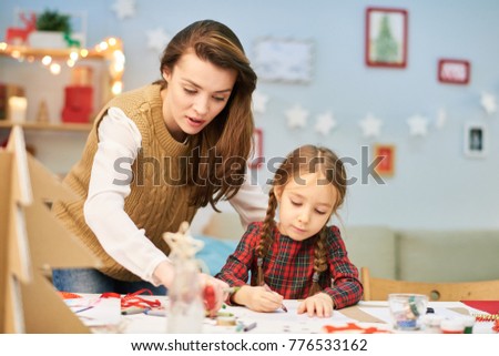 Lovely moments of motherhood: pretty woman wearing knitted vest standing at desk while helping her little daughter to make Christmas card for granny, interior of decorated living room on background