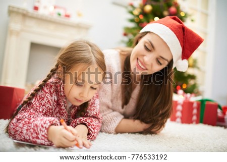 Adorable little girl wrapped up in writing letter to Santa Claus while lying on carpet by fireplace, her attractive mother with charming smile keeping eye on her