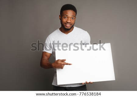 Picture of young smiling african-american man holding white blank board and pointing on it, on grey background, copy space