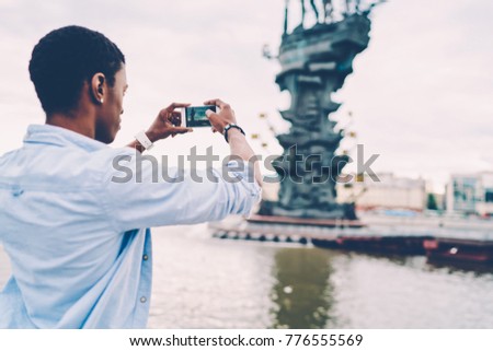 Back view image of male dressed in shirt trying to make good picture of showplace while walking near to river.Afro american man taking photo of landscape on telephone camera during strolling outside