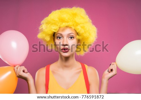 young beautiful girl in yellow clown wig holding multicolored balloons in hands