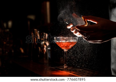 Bartender in black apron and blue shirt sprays an orange peel in cocktail glass with ice at a bar counter Royalty-Free Stock Photo #776607895