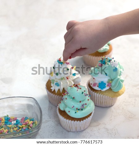A child's hand decorates cooked cupcakes with cream. Bakery products. A square picture.