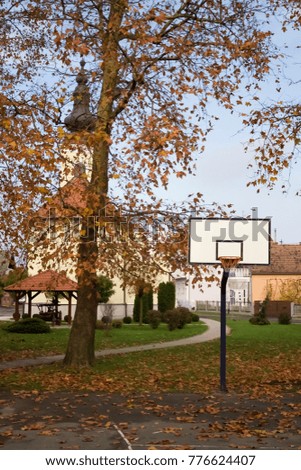Empty basketball court in autumn covered with yellow leaves and church hiding behind tree