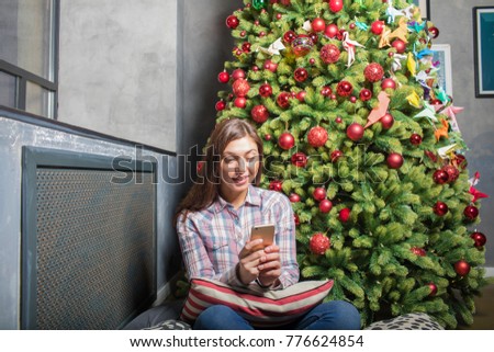 Charming woman received a congratulations with the New Year in text message on mobile phone, sitting near magic Christmas tree with decoration during December holidays. Female chatting on cellphone 