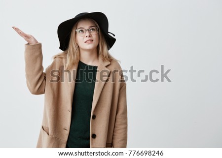 Indignant female student gestures with indignation in coat and black hat, stands in street, doesn`t know what to do because of bad weather. Displeased and dissatisfied blonde girl being puzzled