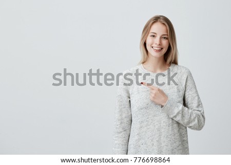 Smiling joyful woman with blonde dyed hair in loose sweater posing against gray studio wall pointing at copy space for advertisment or promotional text. Positive emotions, feelings, joy, happiness