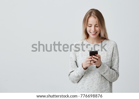 Blonde smiling young woman demonstrating white teeth using cell phone, messaging, being happy to text with her boyfriend, looking at screen of smartphone. Modern technologies and communication