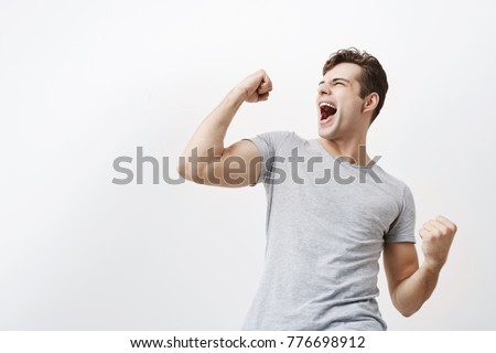 Successful emotional young caucasian male sportsman with dark hair screaming Yes and raising clenched fists in the air, feeling excited. People, success, triumph, victory, winning and celebration. Royalty-Free Stock Photo #776698912