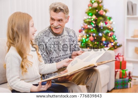 The little girl with a grandfather watch the photo album near the christmas tree