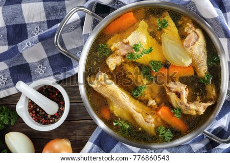 clear Chicken broth with pieces of free range farm organic rooster meat on bone in a metal casserole with kitchen towel and ingredients on wooden table, view from above, close-up