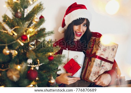 happy stylish woman in santa hat holding many gift boxes with bow at christmas tree lights. space for text. merry christmas and happy new year concept. seasonal greetings, happy holidays
