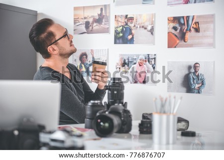 Side view smiling unshaven man watching at different pictures while drinking cup of tea in office. Labor concept
