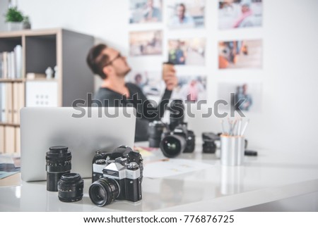 Focus on camera and notebook computer locating on table. Side view serene bearded man looking at photos on wall while tasting mug of coffee. Profession concept