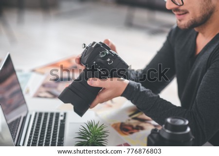 Close up arms of cheerful bearded man holding professional camera. He looking at it while situating at desk with laptop. Job concept