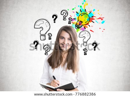 Portrait of a smiling young woman wearing a white blouse and holding an organizer and a pen. A concrete wall background with a colorful lightbulb sketch and many question marks