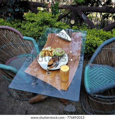 A square picture of a glass and wooden table with two wicker chairs outdoors on summer, with a glass of orange juice and a plate with burger, chips, salad, ketchup and napkins on the table