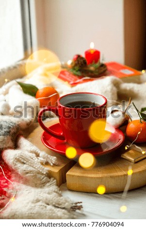 Red cup with hot coffee on the wooden cut with clementine mandarin, candle, book and glowing christmas lights on the window sill. Christmas, holiday morning comfort concept