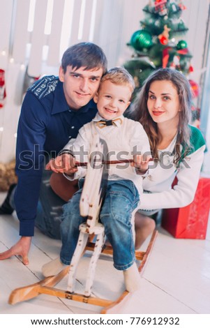 New Year's picture of happy family on background of Christmas decorations. Mother and father play with their son sitting and having fun on the wooden horse