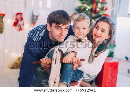 New Year's picture of happy family on background of Christmas decorations. Mother and father play with their son sitting and having fun on the wooden horse