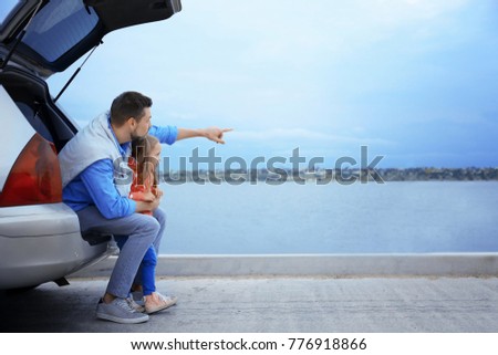 Young father with girl sitting in car trunk near river