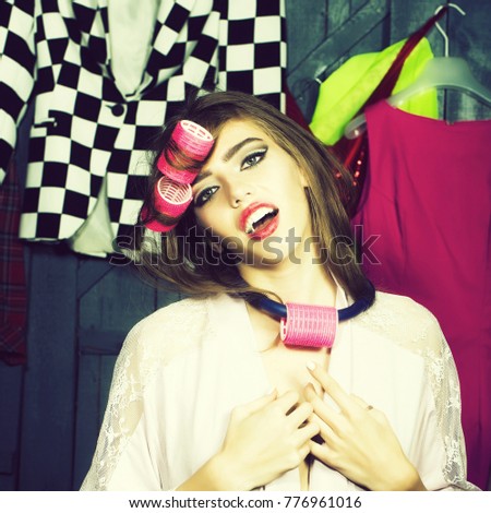 Closeup of one beautiful young fashionable emotional woman in dressing gown with hair-rollers on head standing in wardrobe among many bright clothes, square picture
