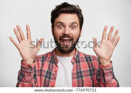Photo of funny bearded male with cheerful expression shows his nonintervention, dressed in checkered shirt, isolated over white background. Happy overjoyed male model gestures actively in studio