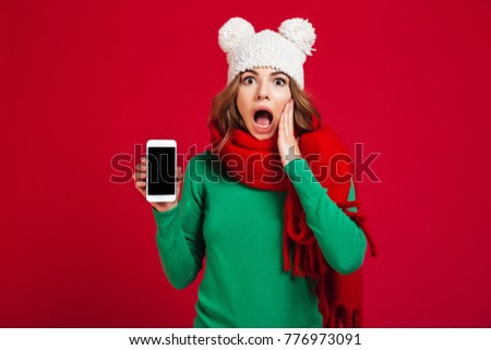 Photo of shocked pretty young woman wearing hat and scarf. Looking camera showing display of mobile phone.