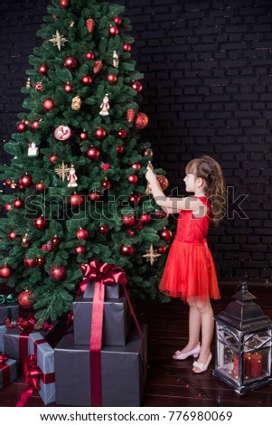 Pretty little girl in red short dress stands near huge Christmas tree decorated with red and golden ornaments for holiday celebration. Many presents under branches. Vertical color photography.