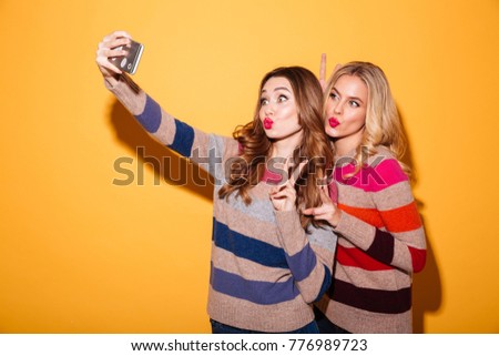 Portrait of two lovely girls dressed in sweaters standing and taking a selfie isolated over white background
