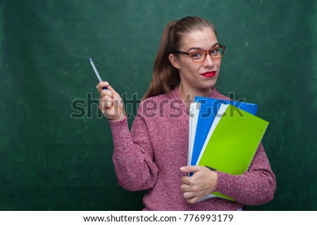Portrait of positive beautiful female student holding hands with training notebooks and ballpoint pen on chalkboard background. Copyspace