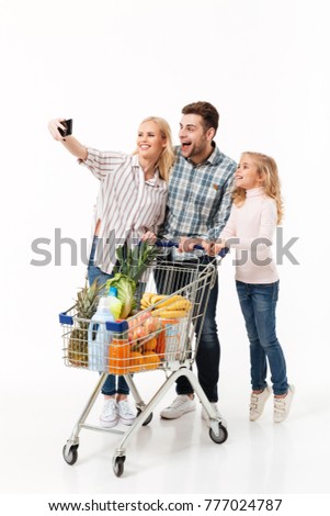 Full length portrait of a happy family standing with a shopping trolley full of groceries and taking a selfie isolated over white background