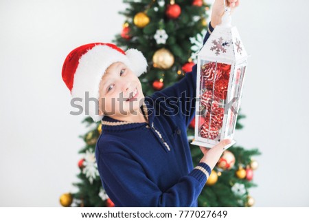 Closeup portrait of cute smiling teenage boy of 10 year old holding winter lantern with red Christmas baubles inside of it. Smiling kid isolated at festive tree background. Horizontal photography.