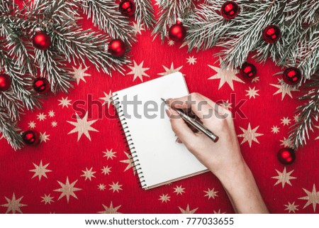 Hands of a girl, woman, writing a Xmas wish text in a notepad, with pen, on a table with red paper stared background, and fir branches view from top, above, Christmas greeting card
