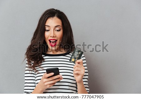 Portrait of an excited girl looking at mobile phone while standing and holding credit card isolated over gray background