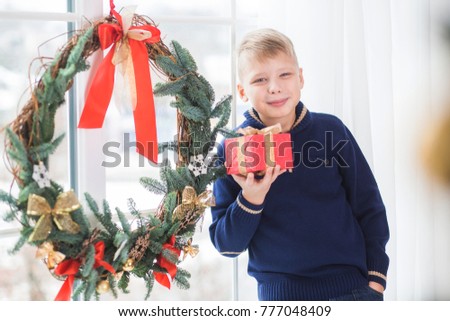 Portrait of cute smiling teenage white boy of 10 year old with present box standing nearl huge Christmas wreath hanging from window. Holiday winter morning concept. Horizontal color photography.