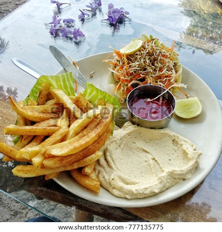 A square picture of a plate with humus, french fries, vegetable salad served in a cabbage leaf, ketchup in a tiny pot, on a table with small purple flowers.