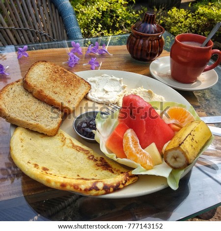 A square picture of a table served with food outdoors on a summer day, surrounded by bushes. There is omelette, two toasts, hummus, tahina, fruit salad and jam on plate. A cup of milk drink and a pot.