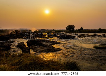 sunset scenery at the victoria falls with some bushland and a unique perspective of the area above the victoria falls with red colors in the sky and rocks between the yellow colored water