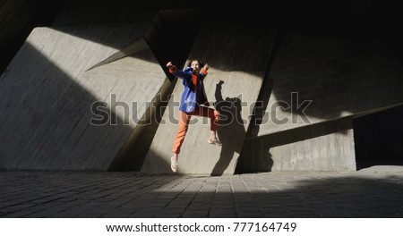 Jumping black-haired woman for joy in violet (purple) T-shirt, orange breeches, a blue-orange leather coat or jacket on background of a bright sunny gray wall with shadows and lights. 